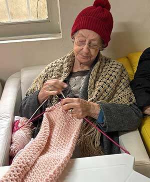 imagem de uma senhora no sofá tricotando uma roupa rosa com uma gorro vermelho na casa de repouso mamma mia imagem de uma senhora no sofá tricotando uma roupa rosa com uma gorro vermelho na casa de repouso mamma mia